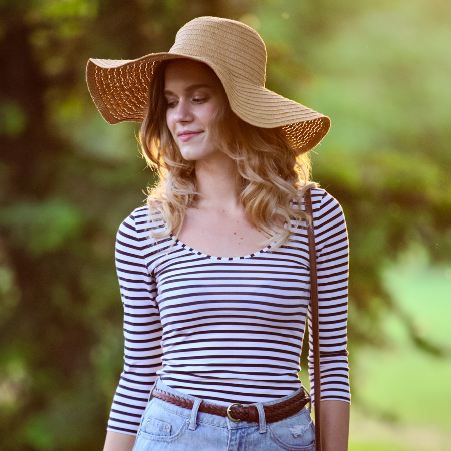 A woman in a hat, striped shirt, and jean shorts walks in a park.